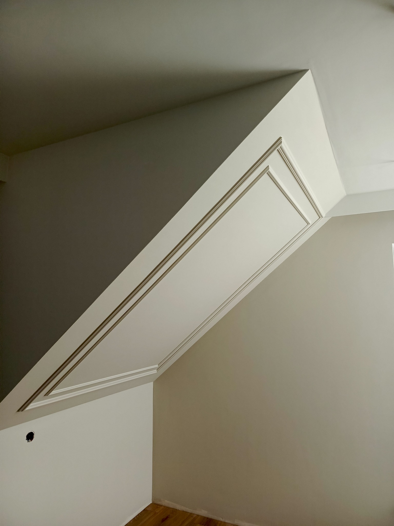 Wykończone poddasze ze skosami, pomalowane na biało z ozdobnymi listwami. Widoczny fragment drewnianej podłogi i otwór na gniazdko elektryczne.