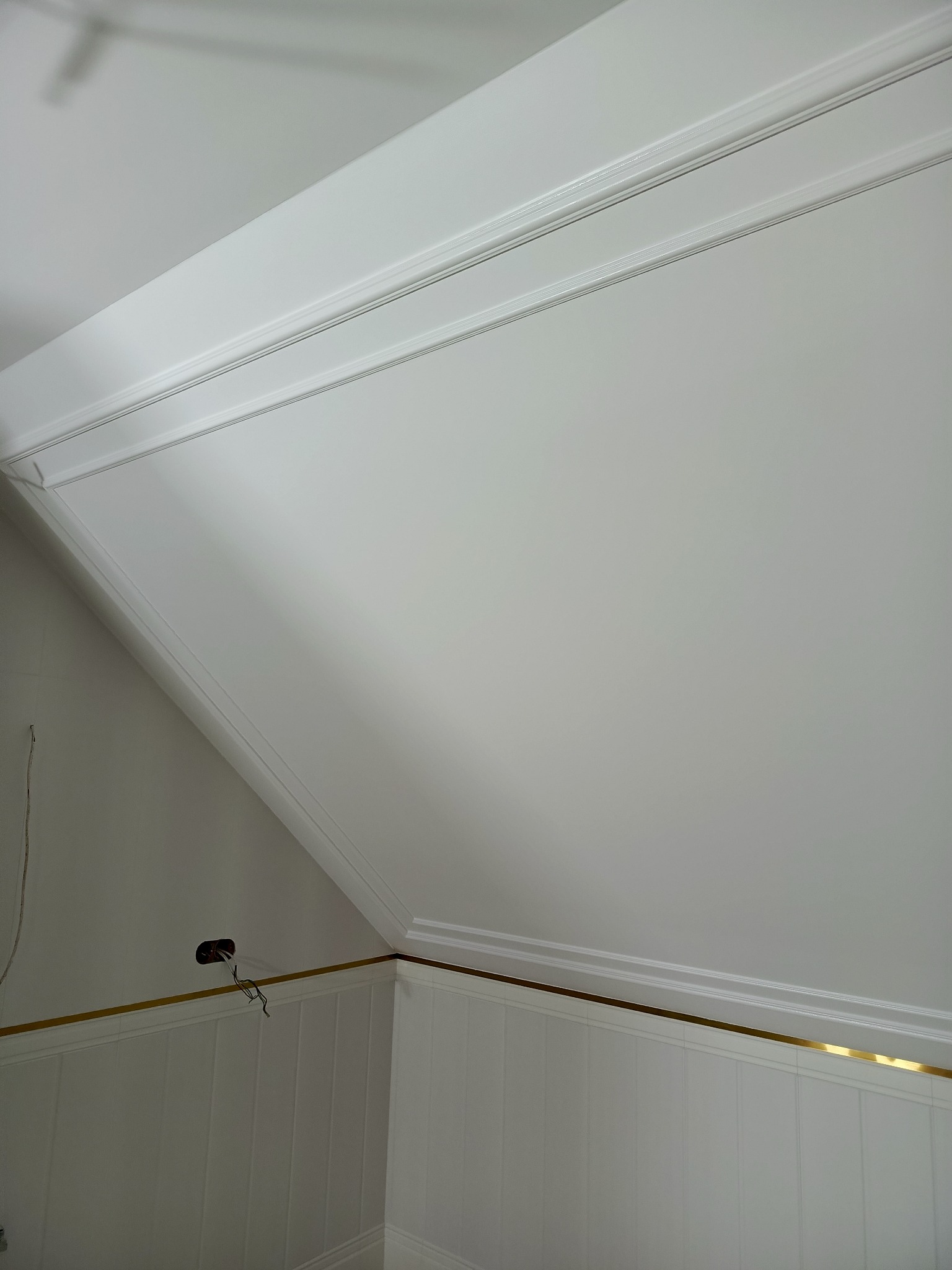 Białe, wykończone panele ścienne z dekoracyjną listwą i gładki sufit pod skosem, ozdobiony sztukaterią. Widoczny fragment instalacji elektrycznej.