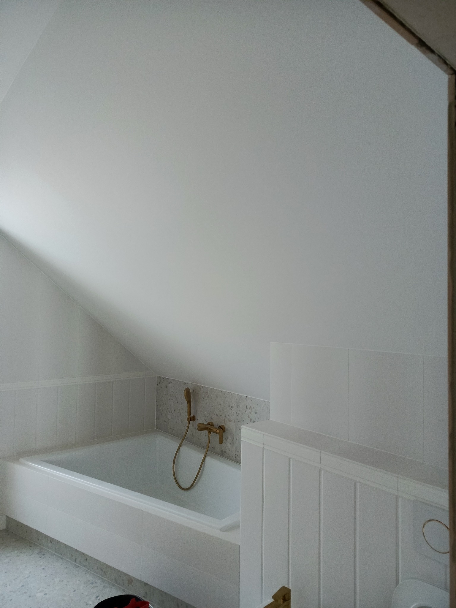 Biała wanna z baterią w kolorze złota w łazience na poddaszu, wykończonej w stylu minimalistycznym z panelami ściennymi i mozaiką nad wanną.