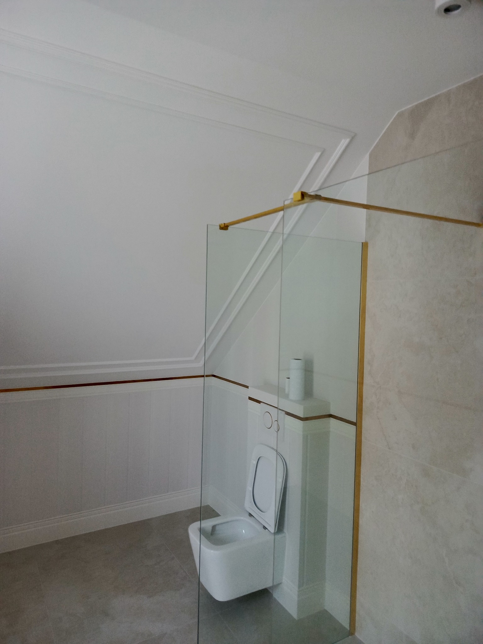 Nowoczesna łazienka na poddaszu z kabiną prysznicową ze złotymi detalami, białą toaletą i szarą podłogą. Wykończenie ścian w bieli i beżu.