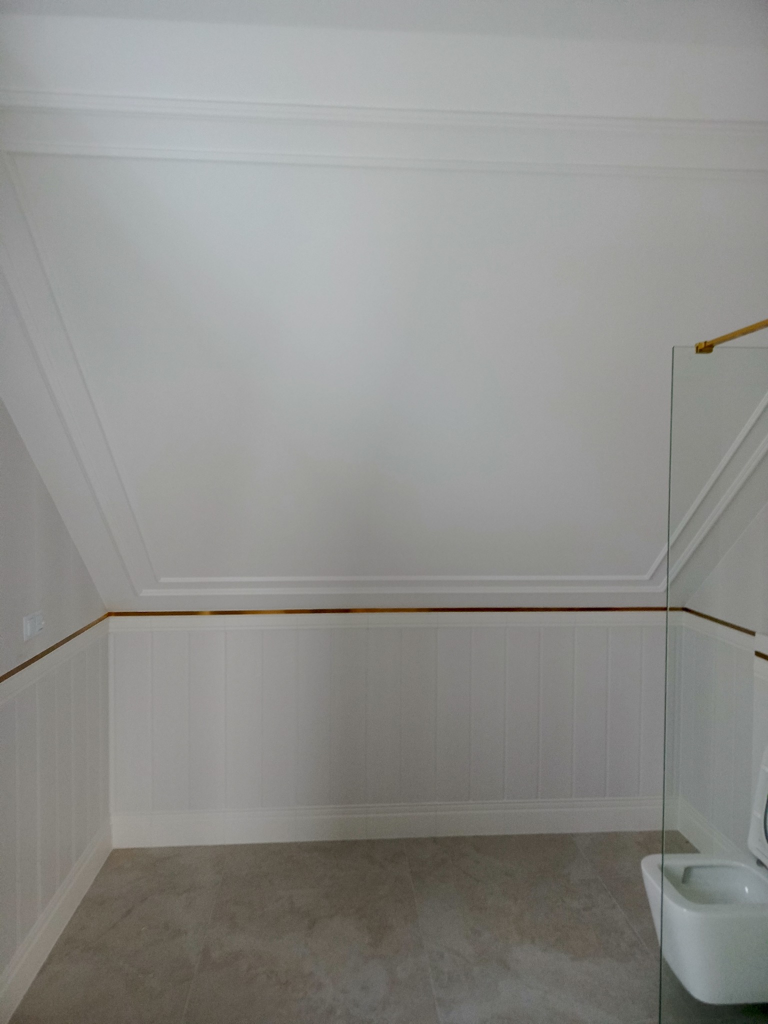 Wykończona łazienka na poddaszu w stylu angielskim: białe panele ścienne z poziomym złotym akcentem, szara podłoga, kabina prysznicowa ze złotymi okuciami, toaleta.