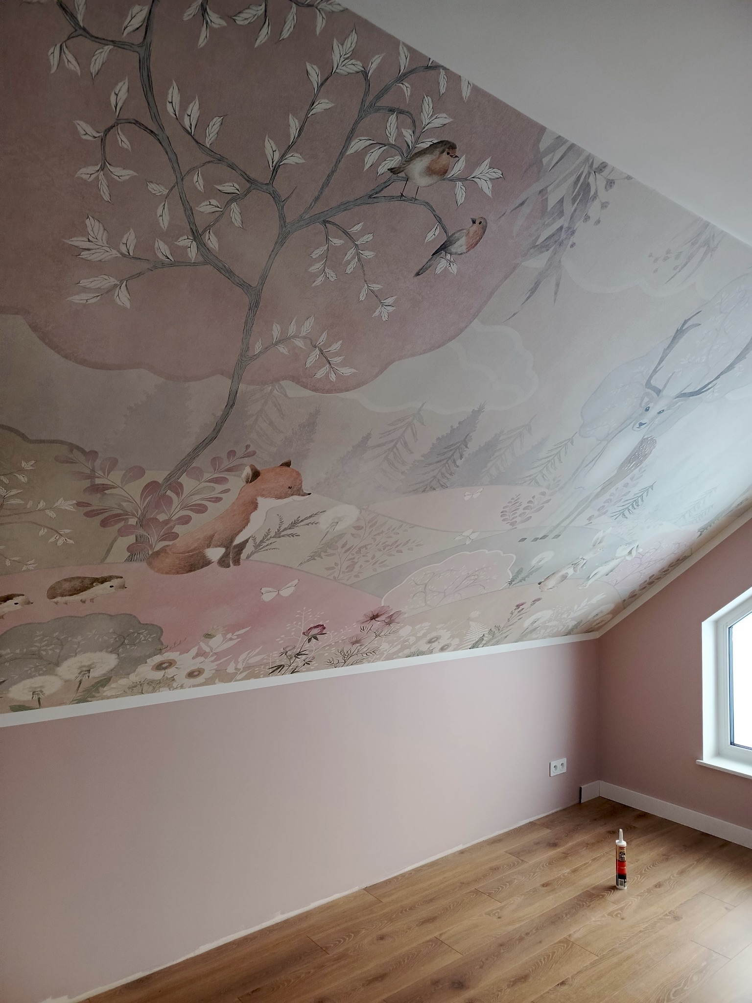 Pokój na poddaszu z ręcznie malowanym motywem leśnym na skośnym suficie, obejmującym drzewa, zwierzęta i delikatne pastelowe kolory. Ściany w odcieniach różu, drewniana podłoga.