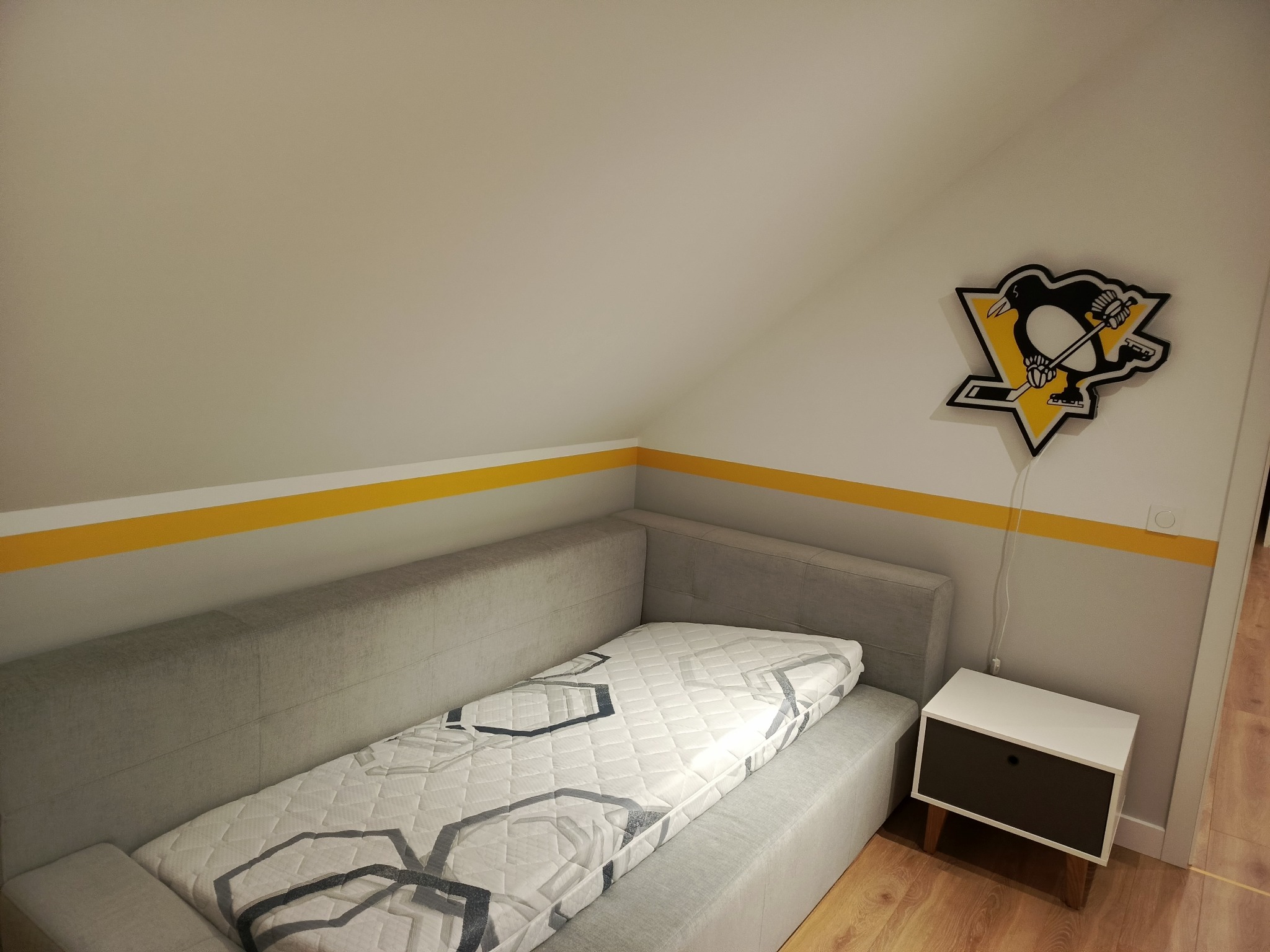 Pokój na poddaszu z szarą sofą i żółtym pasem na ścianie, nad którym wisi lampka z logiem drużyny hokejowej Pittsburgh Penguins. Minimalistyczny styl.