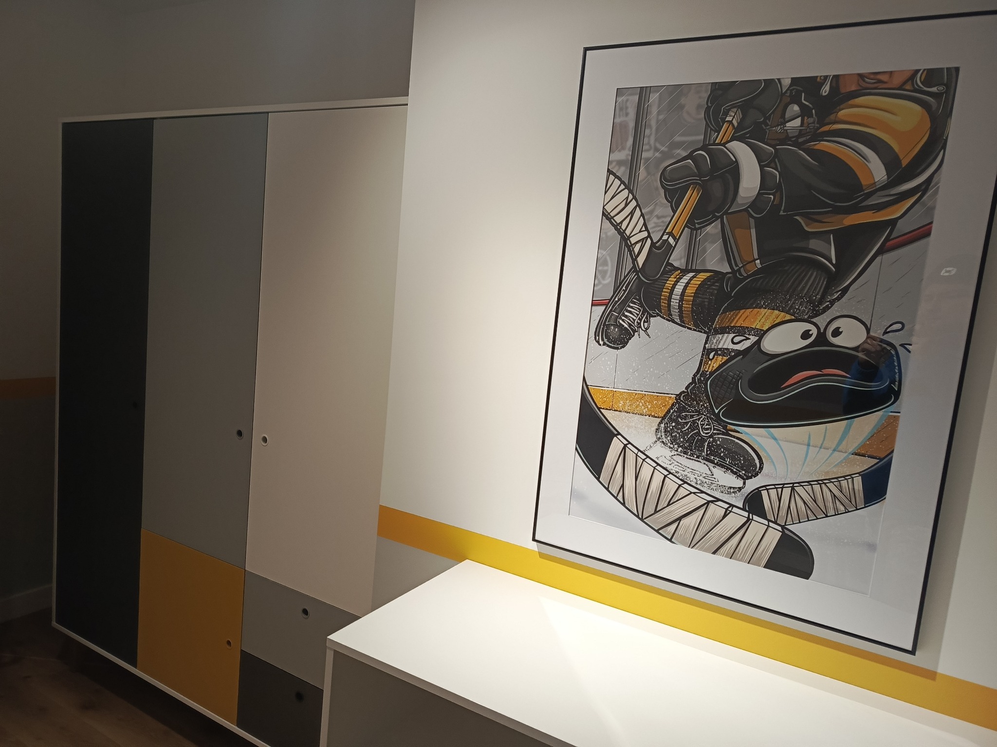 Pokój z szafą w odcieniach szarości i żółci, akcentowany obrazem hokeisty w czarno-żółtej kolorystyce. Nowoczesne wnętrze z minimalistycznym designem.