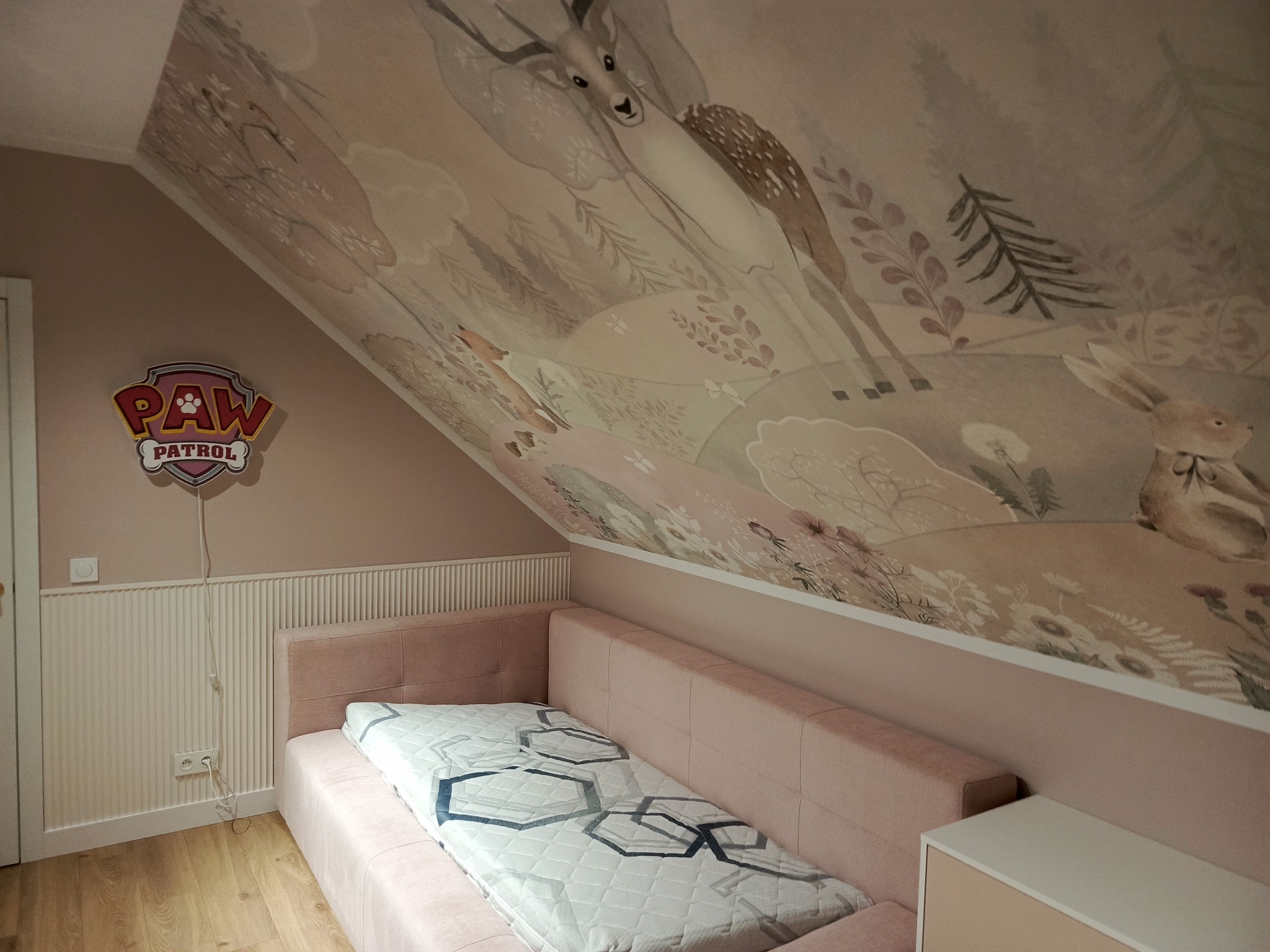 Pokój dziecięcy na poddaszu z łóżkiem i malowidłami zwierząt na skośnym suficie, oświetlony lampką Paw Patrol. Pastelowa kolorystyka.