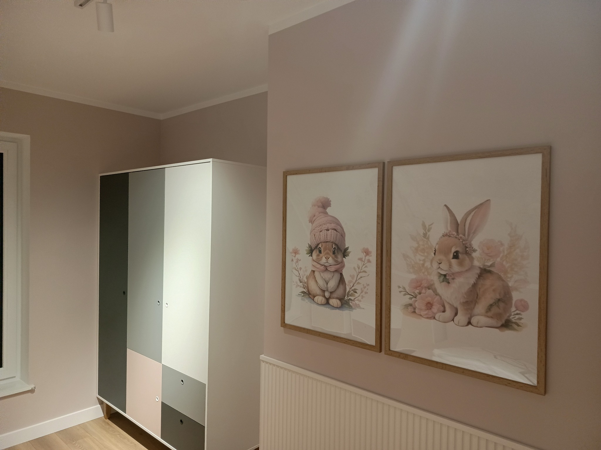 Pastelowy pokój dziecięcy: szafa w odcieniach szarości i różu, dwa obrazy z królikami w drewnianych ramkach na ścianie w kolorze pudrowego różu, biały grzejnik.
