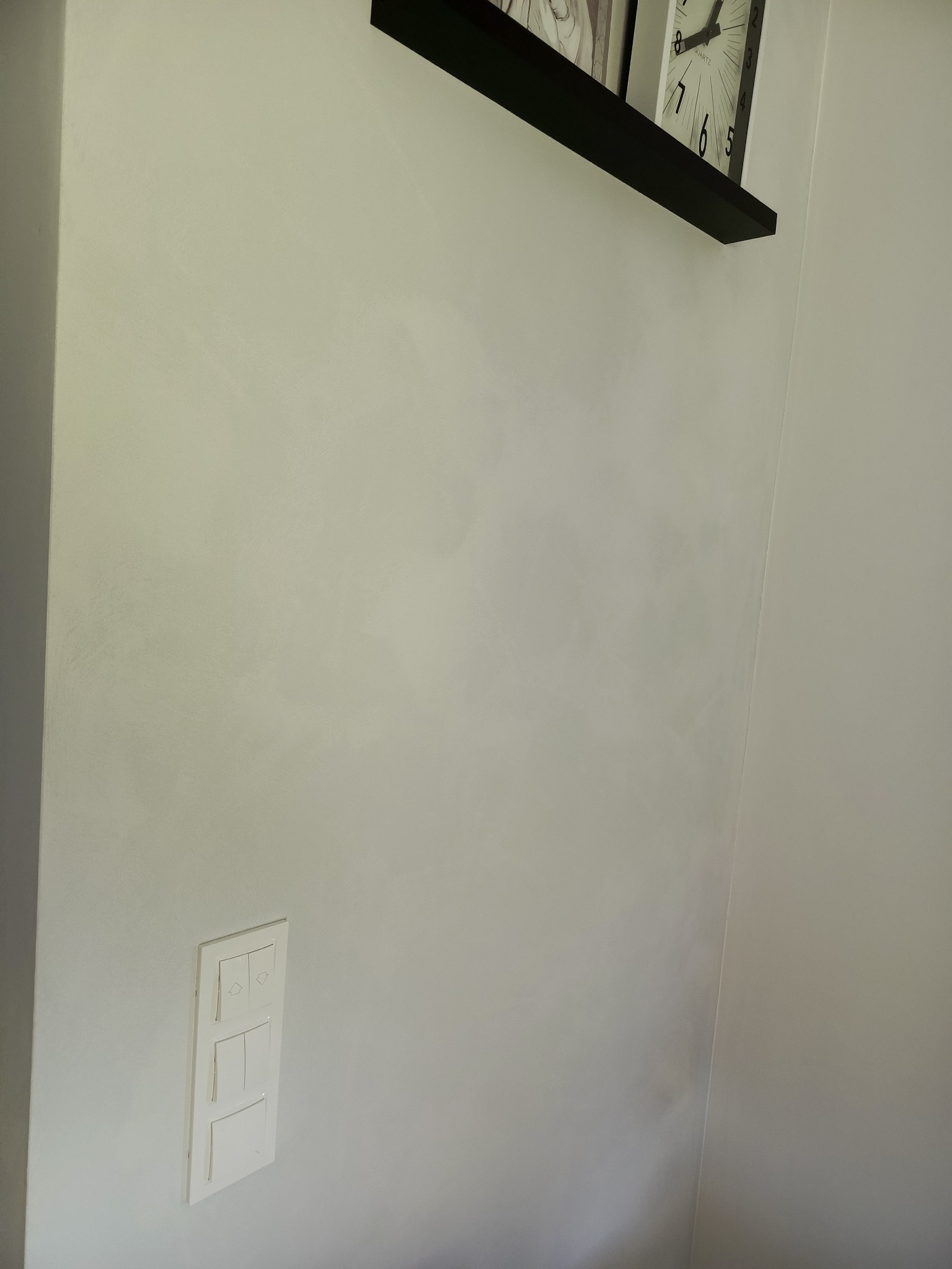 Jasna ściana po szpachlowaniu z widoczną fakturą, w rogu półka z zegarem i zdjęciem, obok włącznik światła. Delikatne oświetlenie podkreśla gładź.