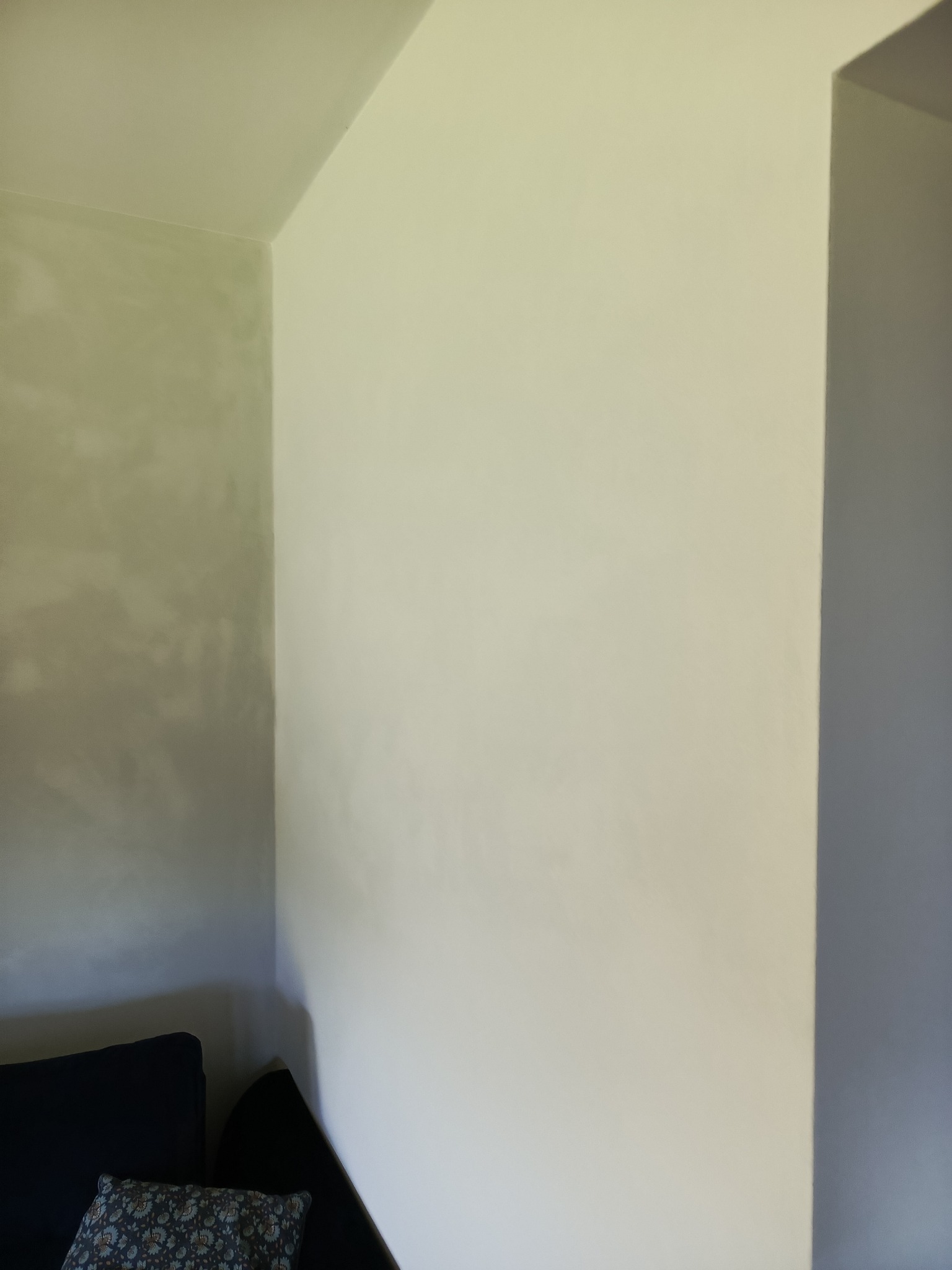 Gładka ściana w odcieniach bieli i szarości, po szpachlowaniu, w rogu pokoju w Siekierczynie. Widoczny fragment sofy z poduszką.