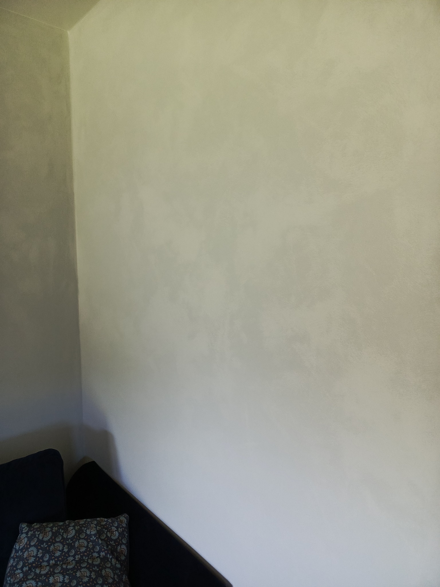 Jasnoszara ściana z efektem dekoracyjnym, widoczna z bliska, z fragmentem sofy w rogu. Delikatna faktura i stonowana kolorystyka.