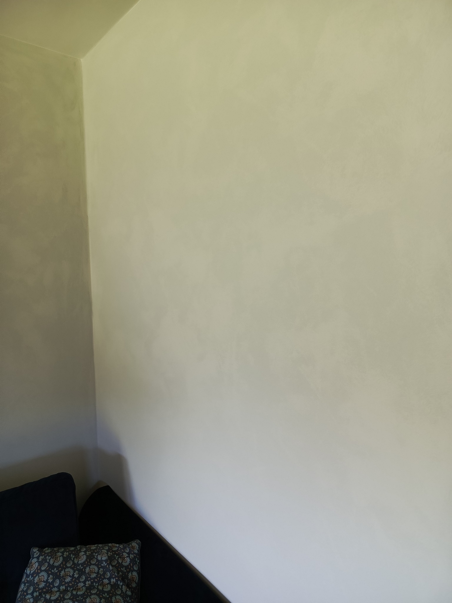 Ściana po szpachlowaniu z efektem dekoracyjnym, widoczna z narożnika pokoju, z fragmentem ciemnej sofy i poduszki w kwiaty.