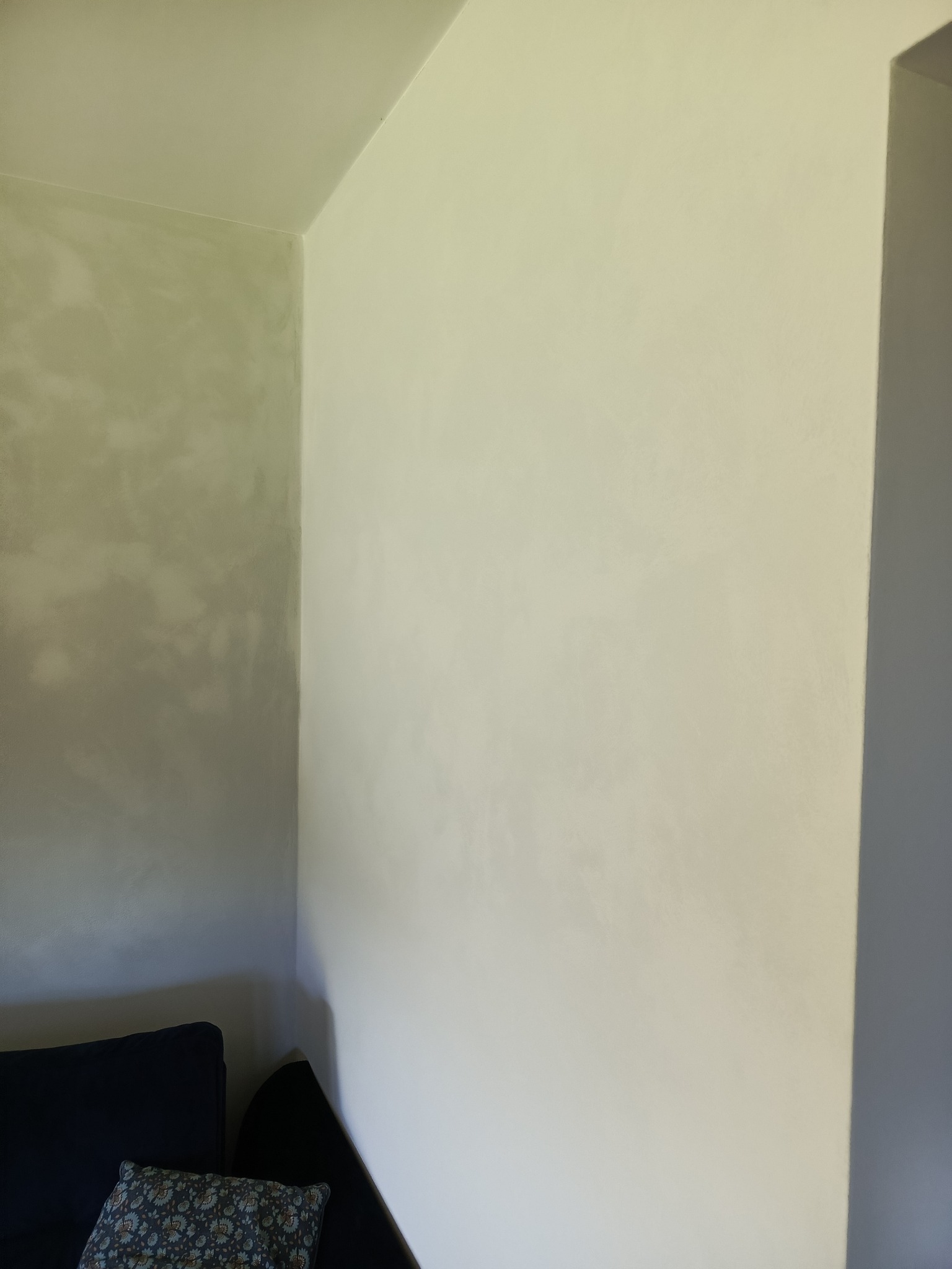 Ściana po szpachlowaniu i malowaniu w odcieniach bieli i szarości, z fragmentem sofy w rogu kadru. Widoczna faktura ściany.