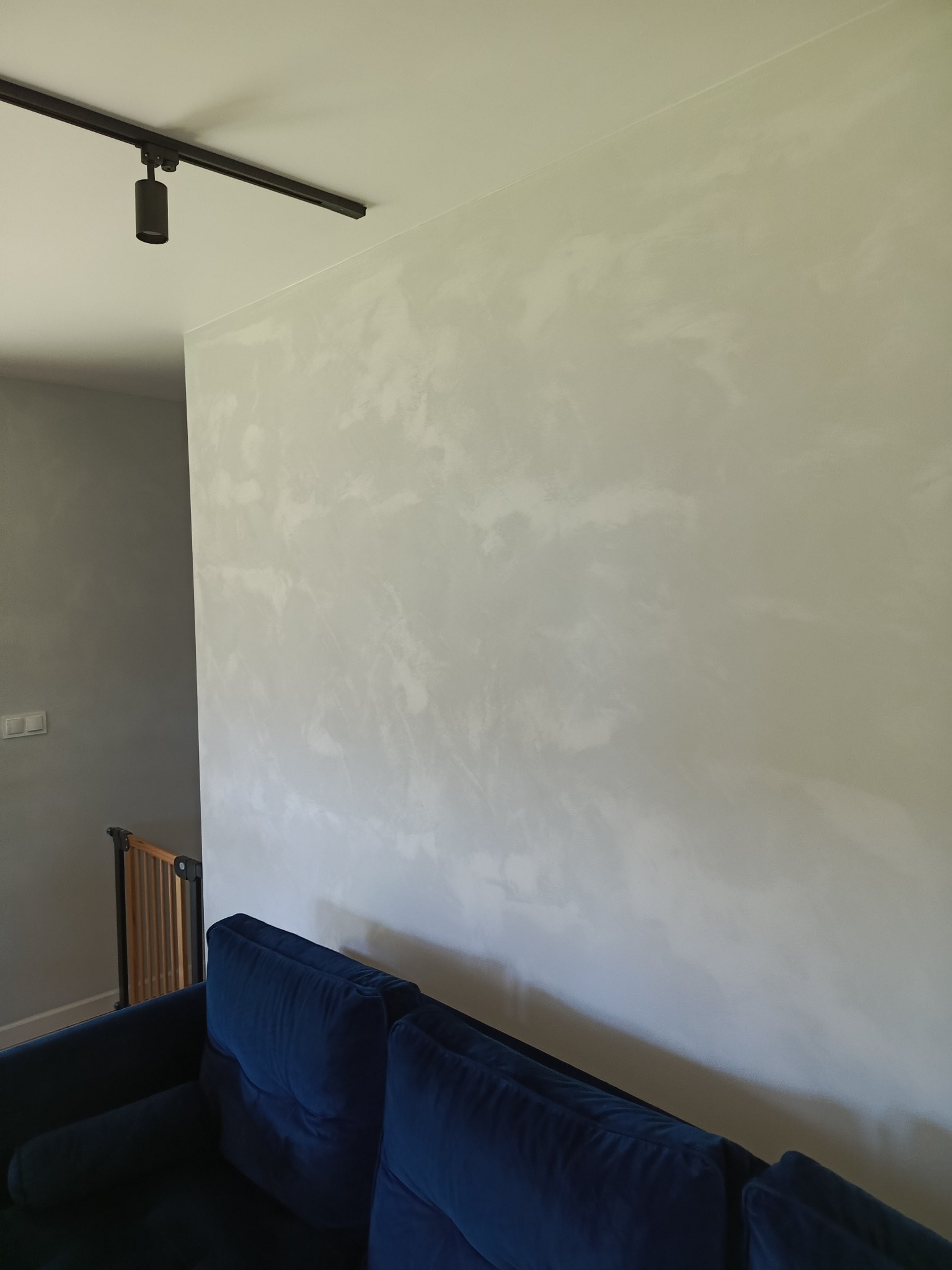 Ściana z efektem dekoracyjnym w odcieniach szarości, oświetlona czarnym reflektorem sufitowym. W tle widoczny fragment niebieskiej sofy i barierka.