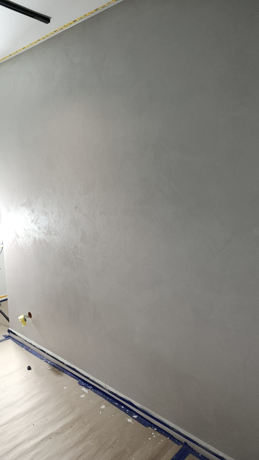 Ściana po gładzi w odcieniach szarości, zabezpieczona podłoga folią malarską. Widoczne delikatne refleksy światła na powierzchni ściany. Gniazdko elektryczne zabezpieczone taśmą.