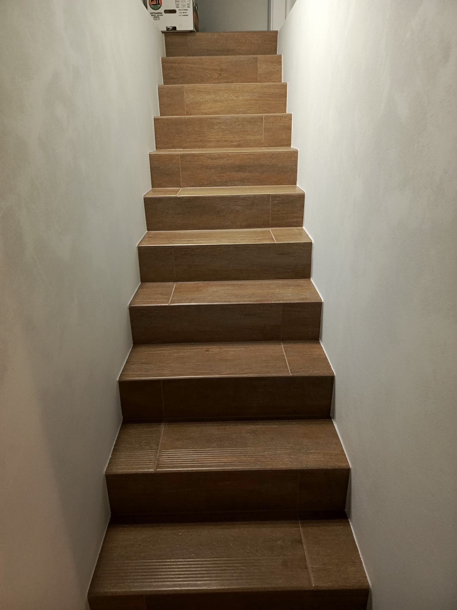 Wykończone schody z płytek imitujących drewno, prowadzące w górę. Ściany pomalowane na jasny, neutralny kolor. Na górze schodów widoczny karton.