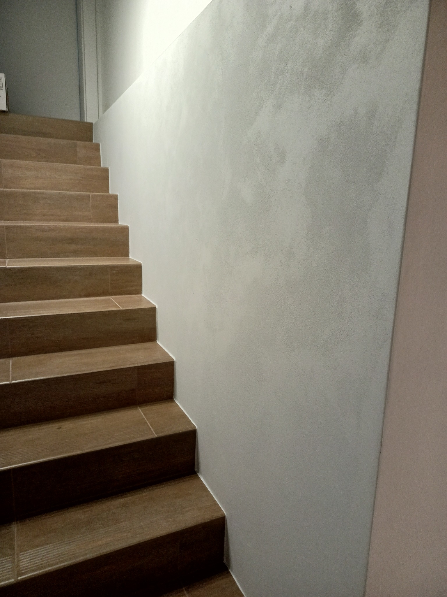 Schody wyłożone brązowymi płytkami imitującymi drewno, przylegające do ściany z szarą, dekoracyjną strukturą. Minimalistyczny design.