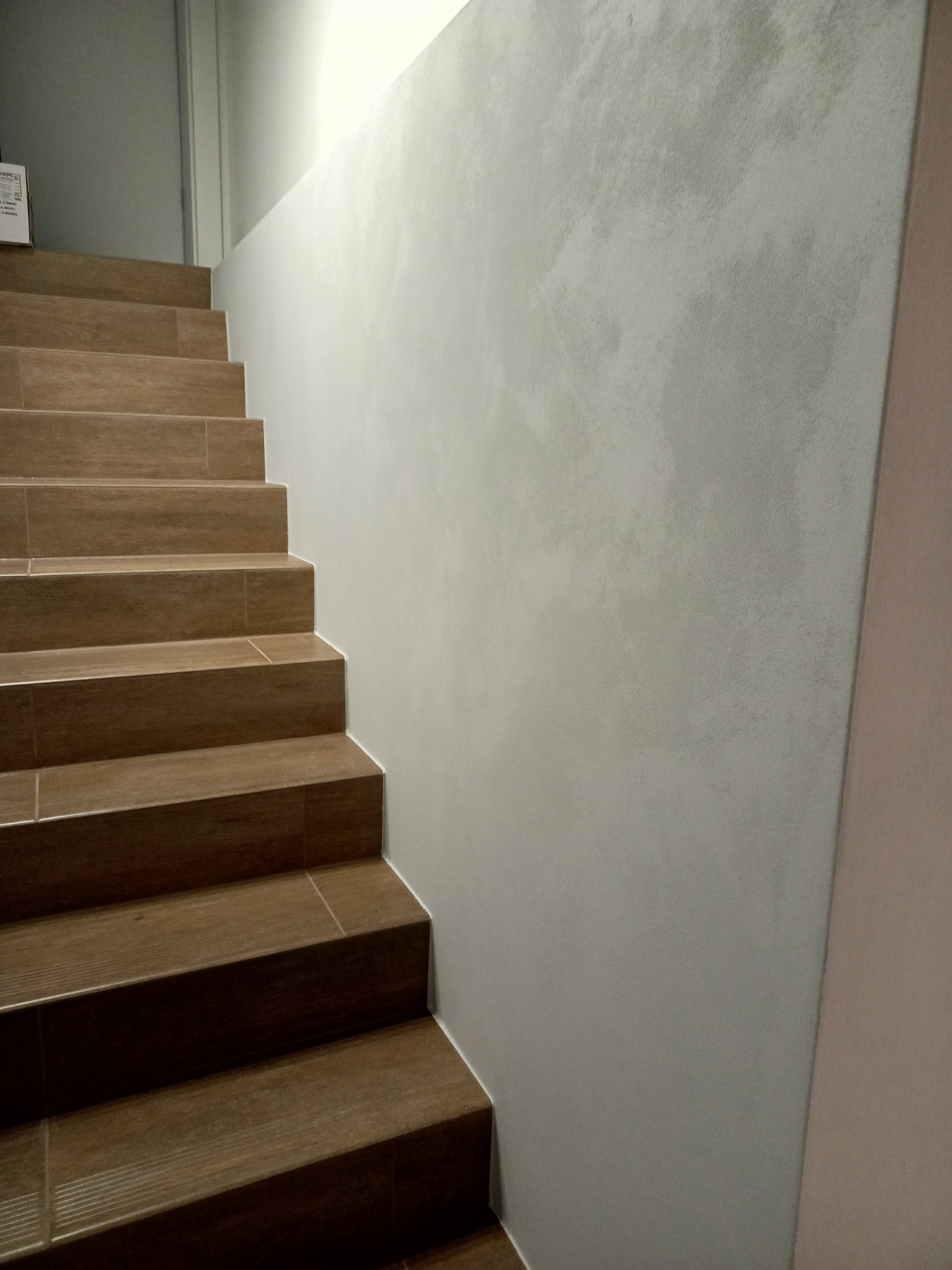 Schody wyłożone brązowymi panelami imitującymi drewno, przylegające do ściany z szarą gładzią. Minimalistyczne wykończenie wnętrza klatki schodowej.
