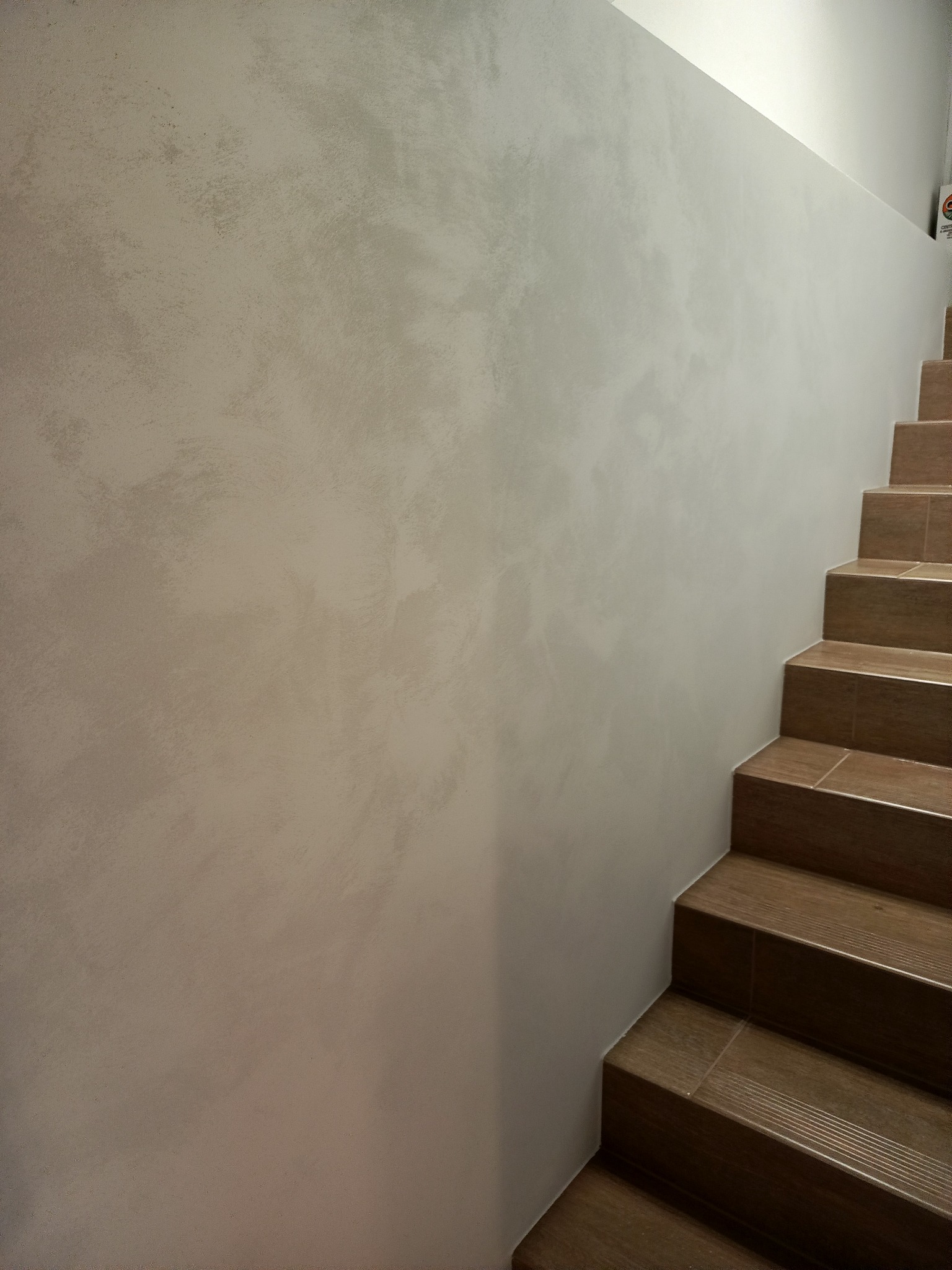 Ściana wewnętrzna z dekoracyjną, szarą gładzią wenecką, sąsiadująca z brązowymi schodami wyłożonymi płytkami imitującymi drewno. Minimalistyczne wykończenie.