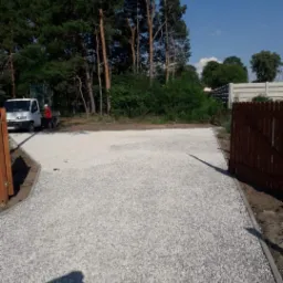 Utwardzony plac z jasnego kruszywa, widoczny biały samochód dostawczy i pracownik w czerwonej koszulce, w tle drzewa i ogrodzenie.