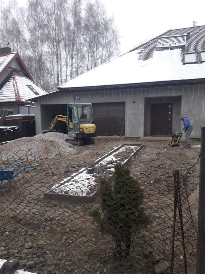 Prace ziemne na posesji: minikoparka obok sterty gruzu, pracownik zagęszcza teren ubijarką, widoczny zarys fundamentów pod obrzeże trawnika, zimowa sceneria.
