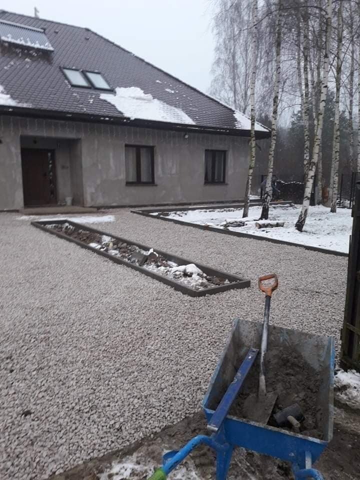 Podjazd z jasnego żwiru przed szarym domem z brązowym dachem, w tle brzozy, niebieska taczka z narzędziami i zaprawą na pierwszym planie w zimowej scenerii.