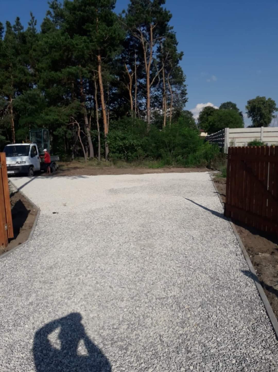 Utwardzony plac z jasnego kruszywa, widoczny biały samochód dostawczy i pracownik w czerwonej koszulce, w tle drzewa i ogrodzenie.