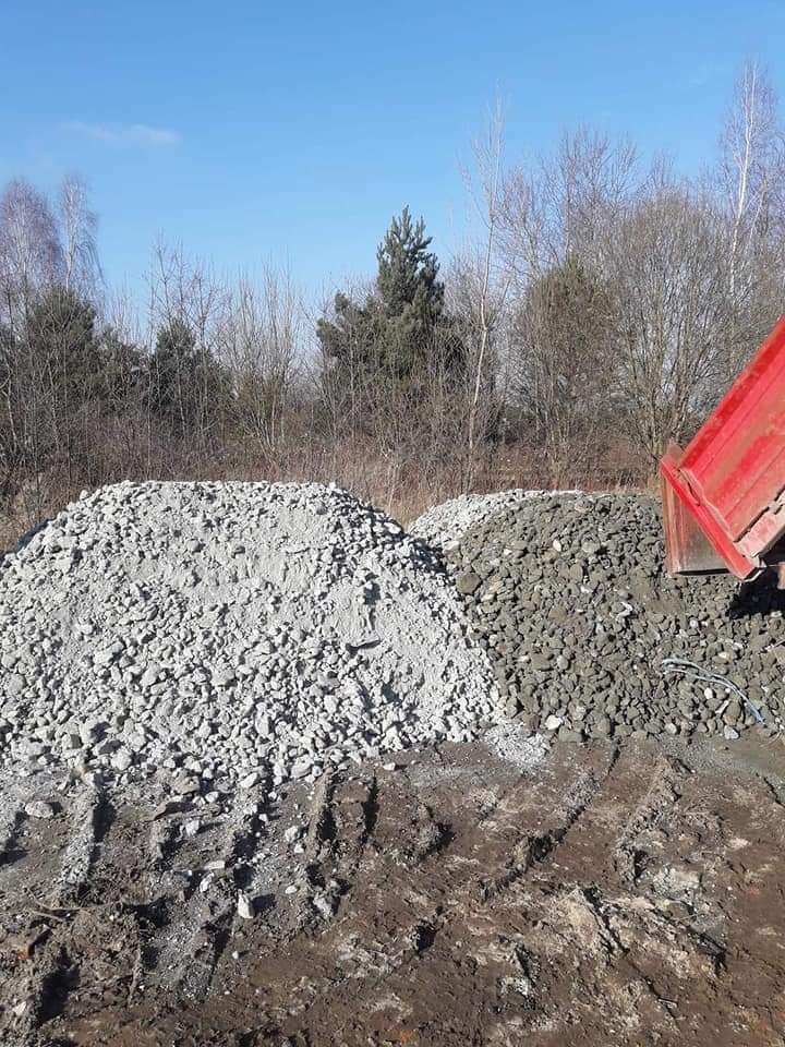 Dwie pryzmy: jasnego i ciemnego kruszywa, świeżo wysypane z wywrotki na placu budowy, z widocznymi śladami opon w ziemi i fragmentem czerwonej burty samochodu ciężarowego.