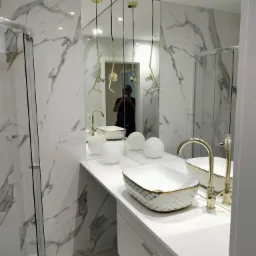 Elegancka łazienka z marmurowymi płytkami, złotą armaturą i dekoracyjną umywalką. Lustro powiększa przestrzeń, a nowoczesne lampy dodają blasku.