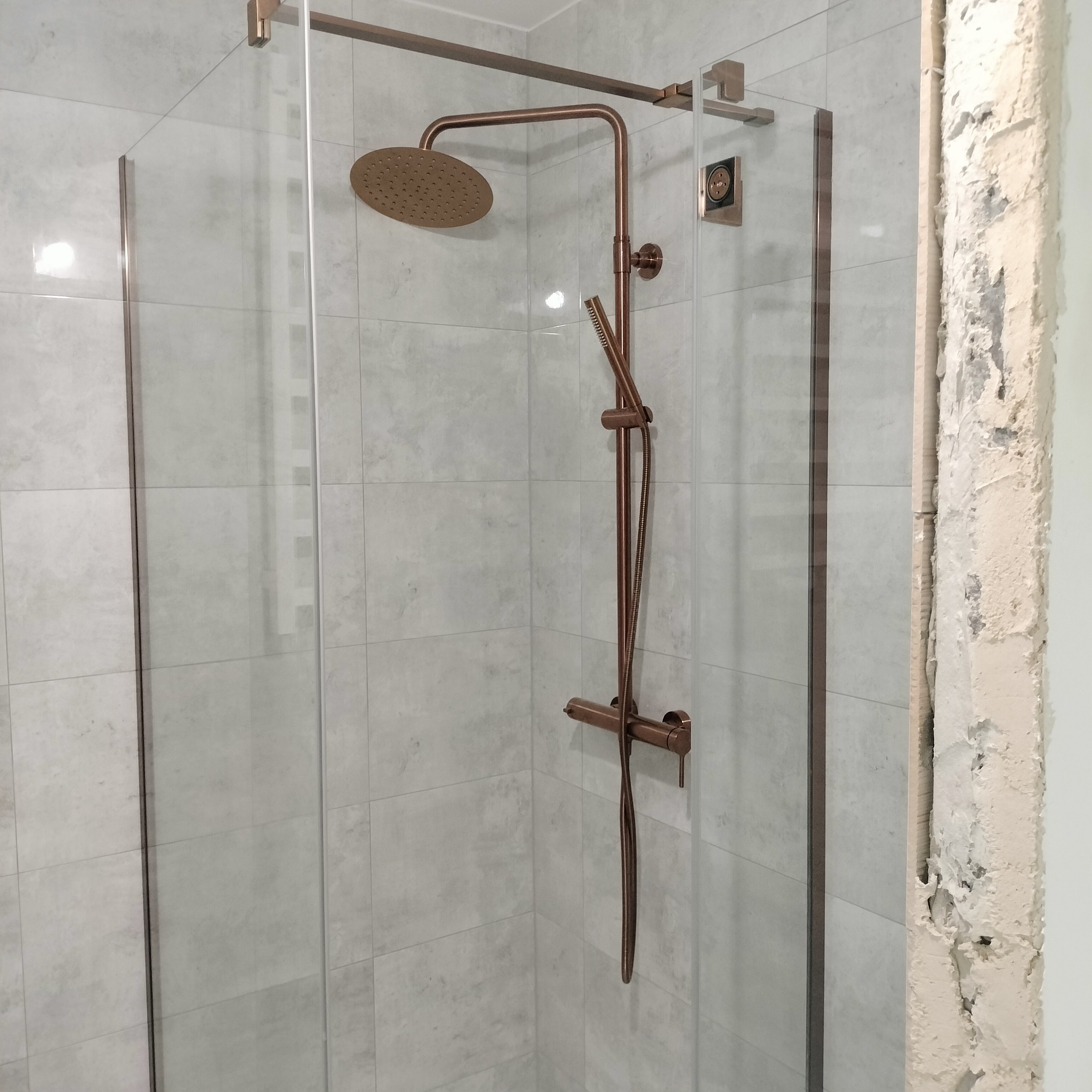 Kabina prysznicowa z armaturą w odcieniach miedzi na tle jasnych płytek. Fragment ściany w stanie surowym. Widok na wykończoną część łazienki w trakcie prac.