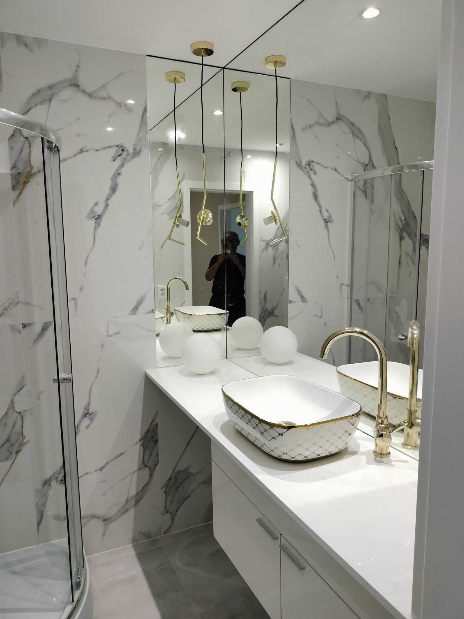 Elegancka łazienka z marmurowymi płytkami, złotą armaturą i dekoracyjną umywalką. Lustro powiększa przestrzeń, a nowoczesne lampy dodają blasku.