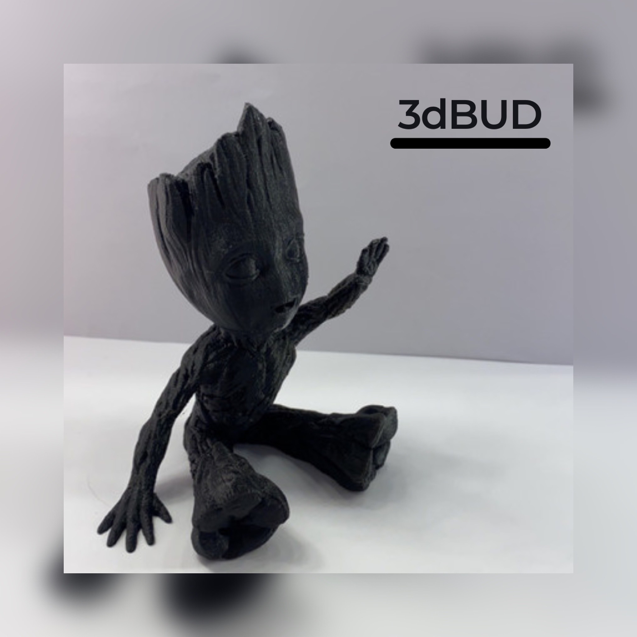 Czarna figurka Baby Groot wydrukowana w 3D, siedząca na białym tle, z widocznym logo firmy 3dBUD w tle.