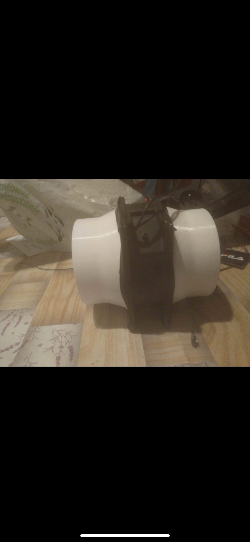 Czarna kabura na białym cylindrze, leżąca na podłodze z wzorem drewna, z widoczną torbą w tle.