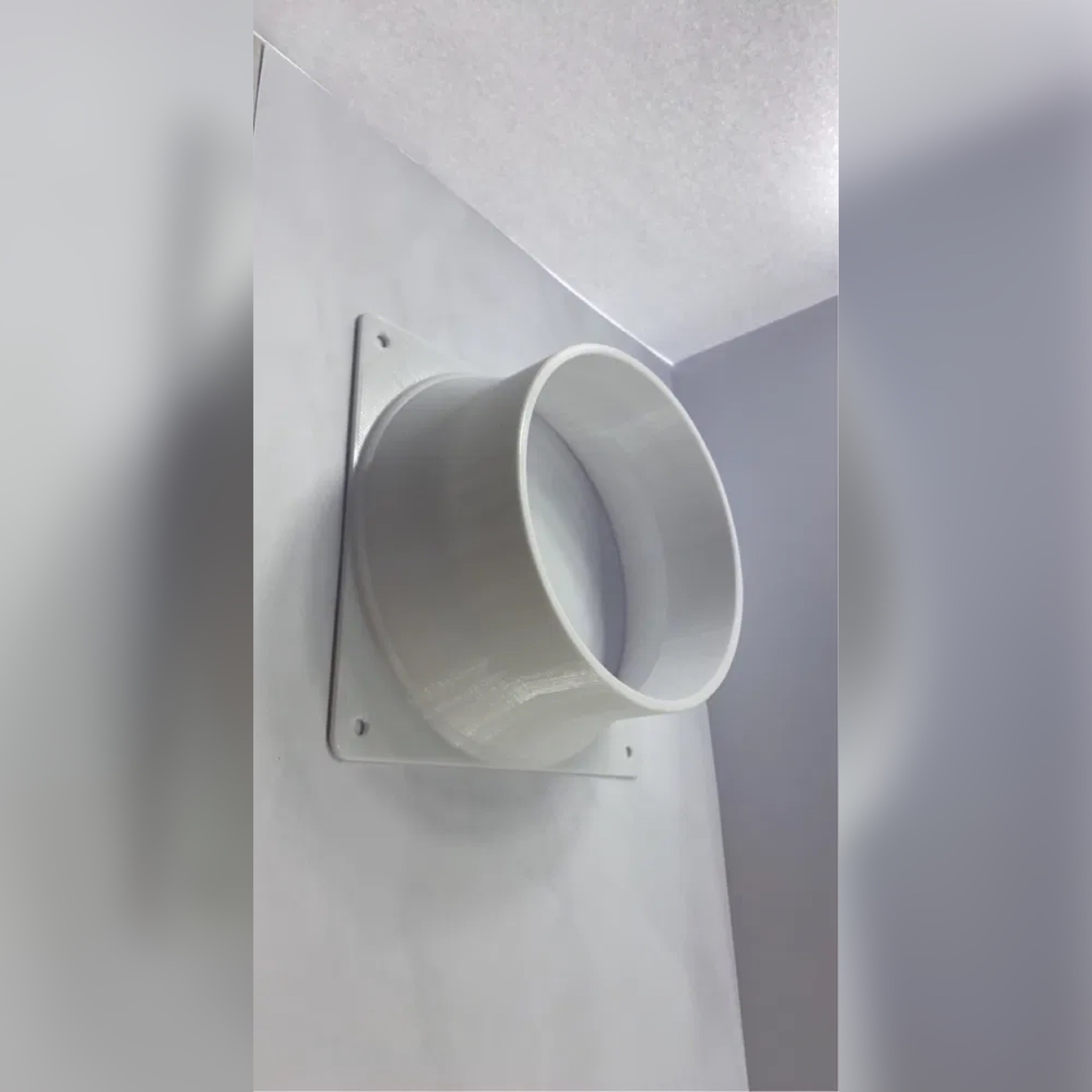 Biały, plastikowy element wentylacyjny zamontowany na jasnej ścianie, widoczne cztery śruby mocujące.