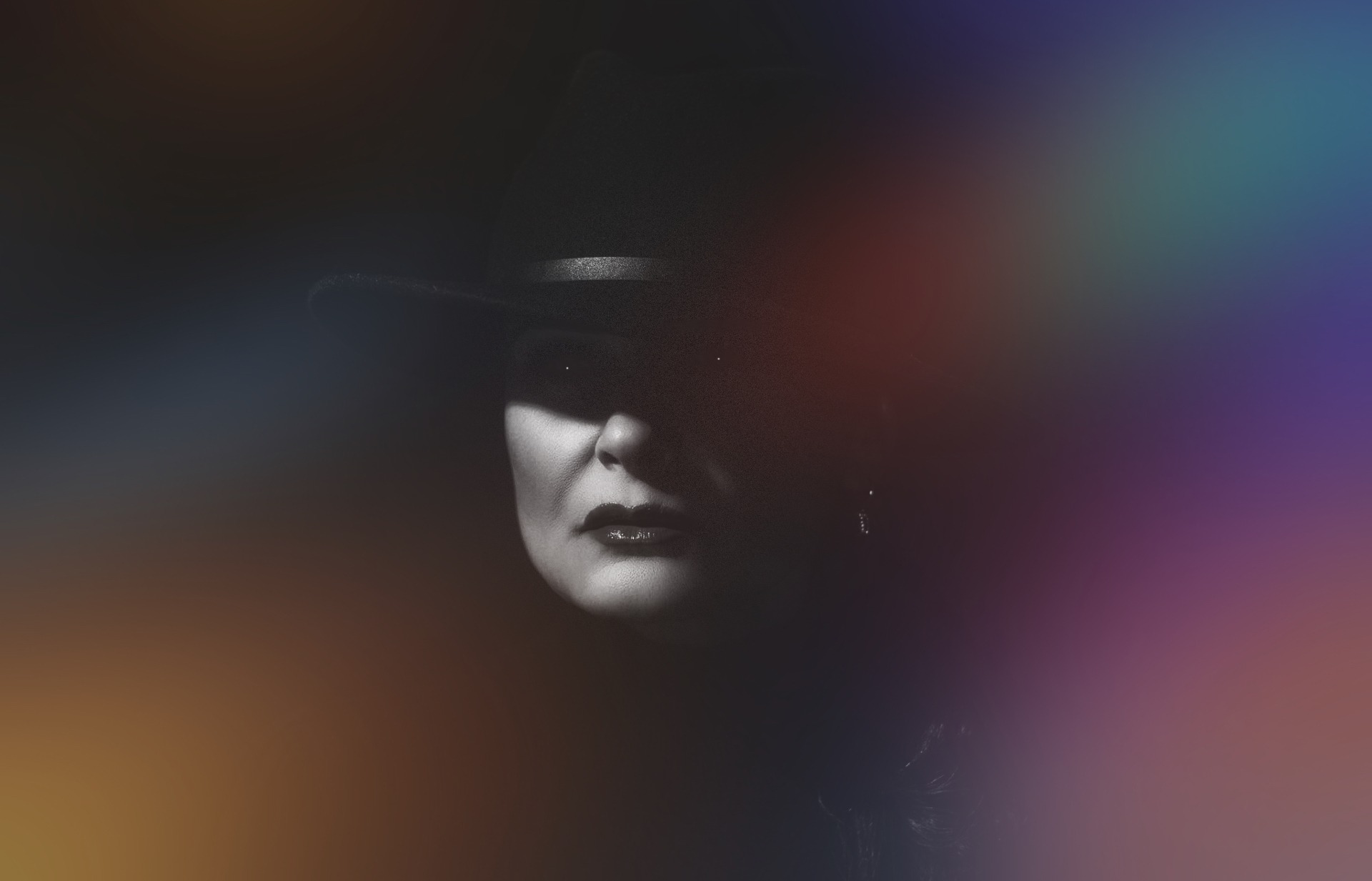 Czarno-biały portret kobiety w kapeluszu z ciemnym tłem i abstrakcyjnymi kolorowymi plamami na obrzeżach.