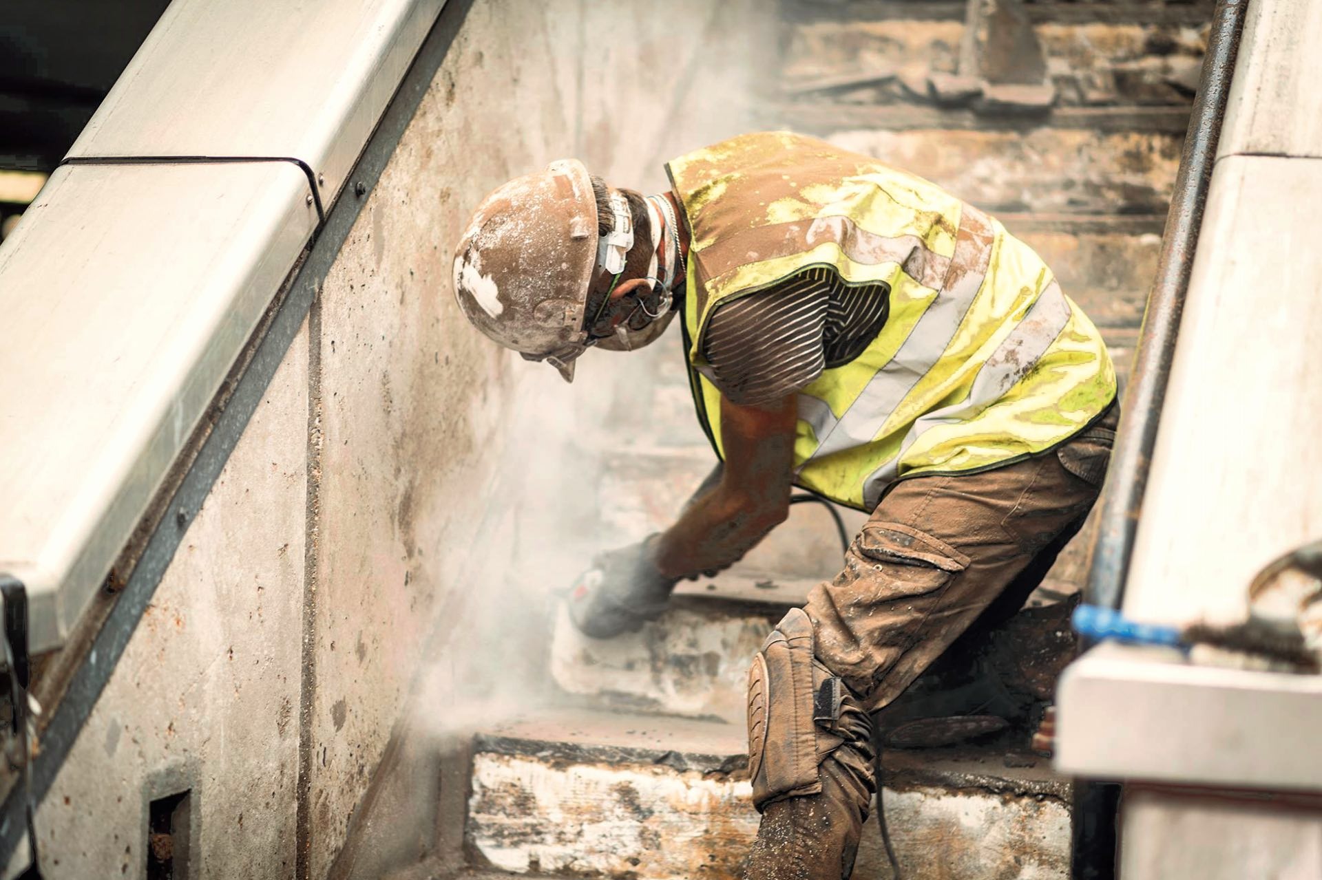 Pracownik w kasku i kamizelce odblaskowej szlifuje betonowe schody podczas remontu, unoszący się pył.