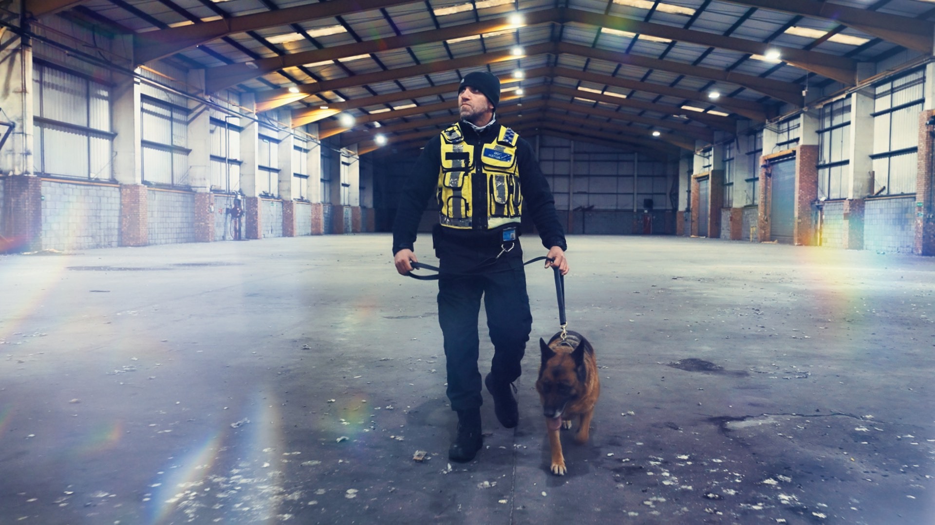 Mężczyzna w kamizelce ochronnej prowadzi owczarka niemieckiego wewnątrz pustego, przestronnego magazynu z betonową podłogą i metalową konstrukcją dachu.