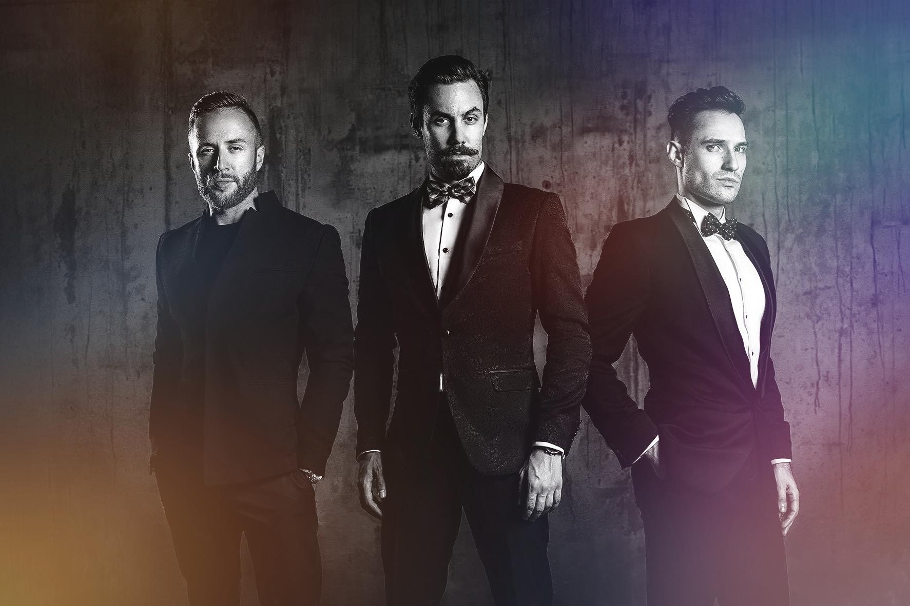 Trzech eleganckich mężczyzn w smokingach i muchach pozuje na tle betonowej ściany z gradientem kolorów.