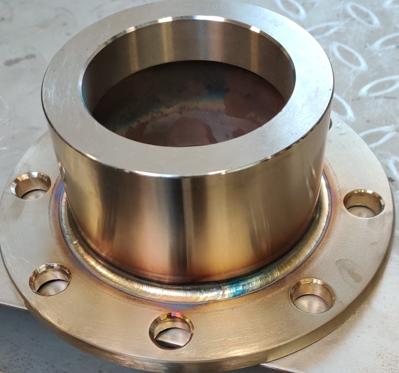 Mosiężny element z kołnierzem i otworami, połączony spoiną z widocznymi śladami ciepła, leżący na metalowej powierzchni z wzorem rombów.