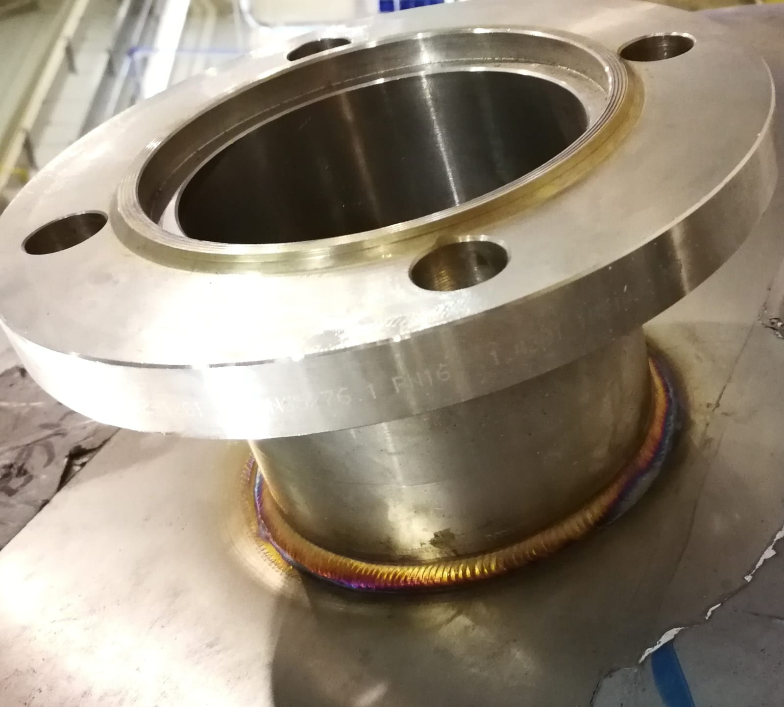 Metalowy kołnierz z widocznym spoiną spawalniczą o tęczowych barwach, przymocowany do płaskiej powierzchni. Widoczne oznaczenia na kołnierzu: NGSx76.1 PN16 1.4301.