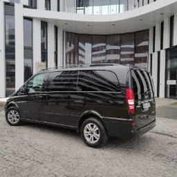 Czarny van Mercedes Viano zaparkowany na brukowanym placu przed nowoczesnym budynkiem biurowym z białymi i czarnymi elementami fasady, tablica rejestracyjna widoczna.