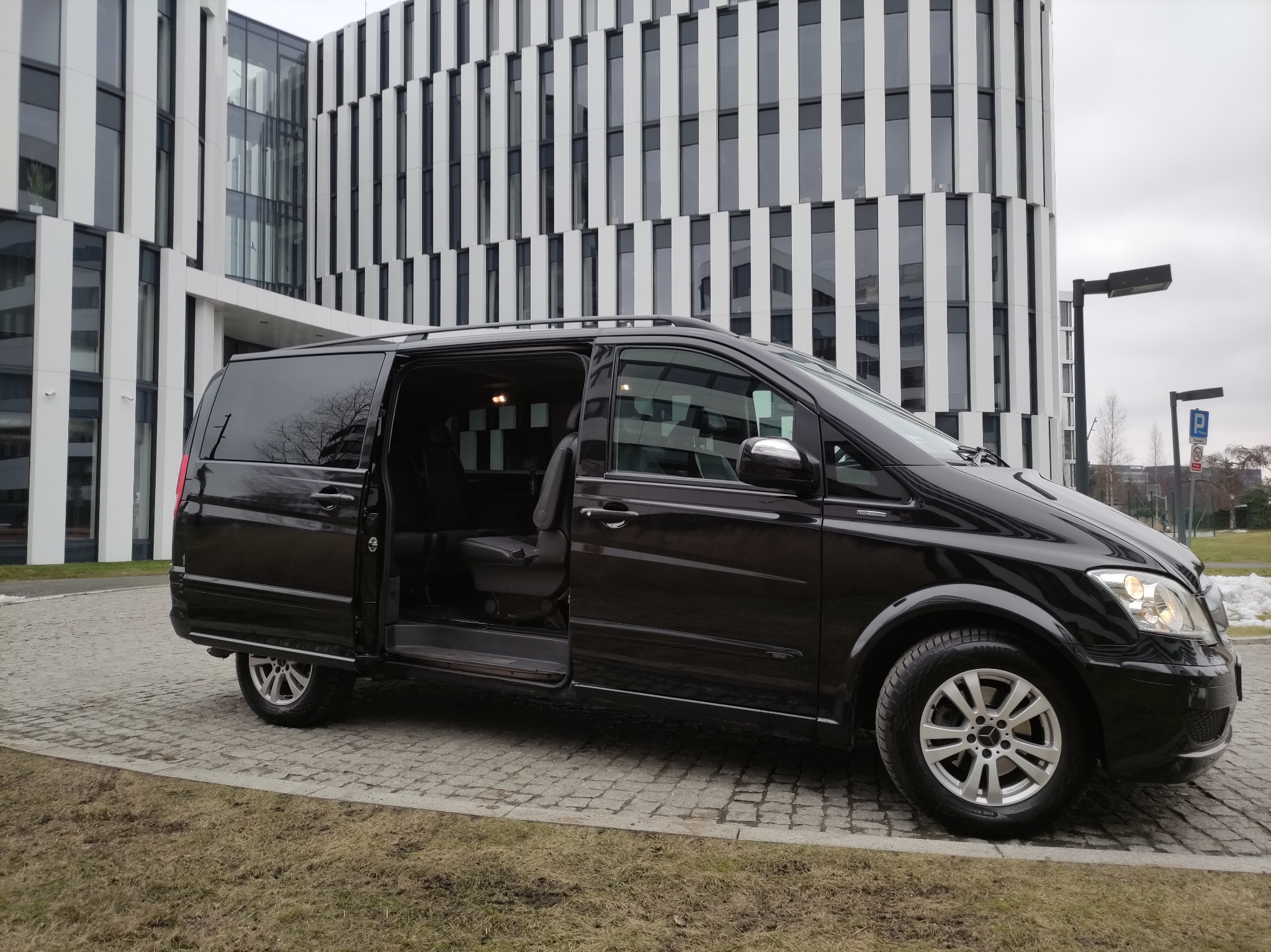 Czarny van Mercedes Viano z otwartymi drzwiami przesuwnymi, zaparkowany przed nowoczesnym budynkiem biurowym z fasadą w biało-czarne pasy.