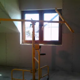 Wnętrze poddasza z oknem, ściany wykończone płytami gipsowo-kartonowymi, widoczny podnośnik do płyt w kolorze ż&oacute;łtym.