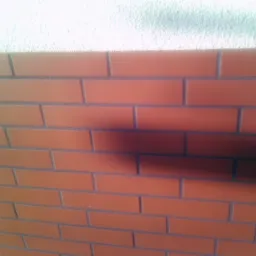 Elewacja budynku wykończona czerwoną cegłą klinkierową z szarymi fugami, górna część ściany pokryta jasnym tynkiem strukturalnym, cień na ścianie.