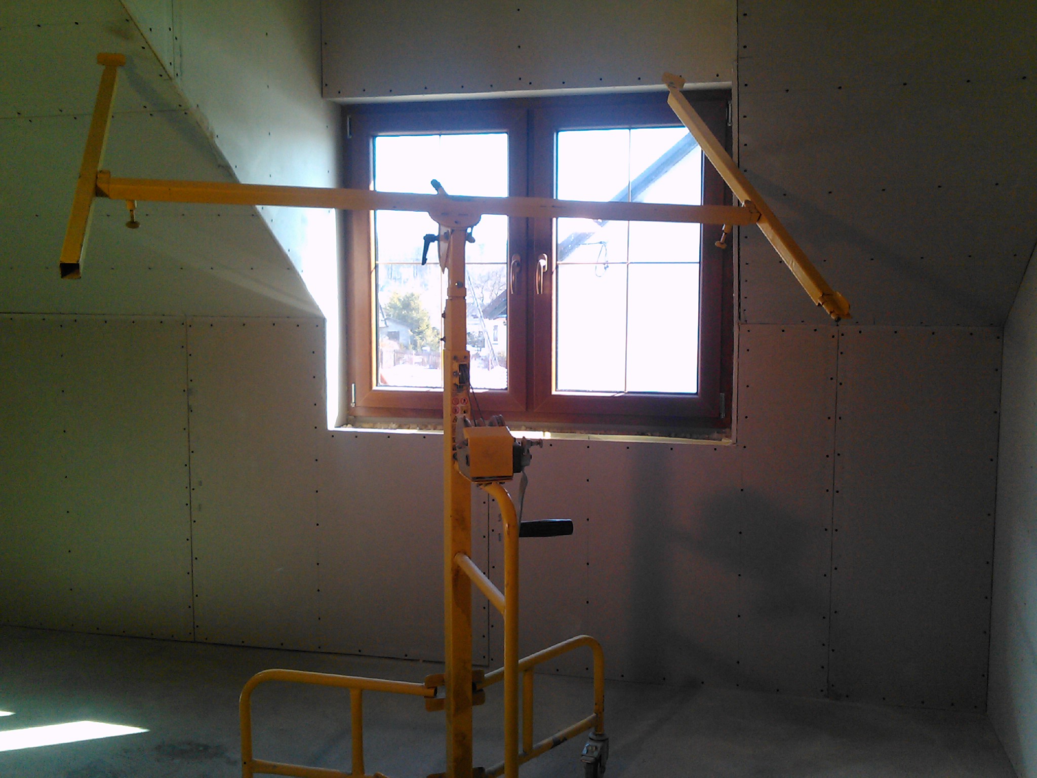 Wnętrze poddasza z oknem, ściany wykończone płytami gipsowo-kartonowymi, widoczny podnośnik do płyt w kolorze żółtym.