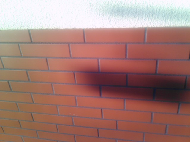 Elewacja budynku wykończona czerwoną cegłą klinkierową z szarymi fugami, górna część ściany pokryta jasnym tynkiem strukturalnym, cień na ścianie.