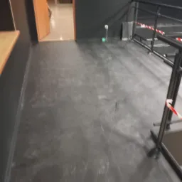 Szara, gładka podłoga w pomieszczeniu z czarnymi ścianami, widoczne fragmenty okna i schodów z barierką zabezpieczoną taśmą ostrzegawczą.