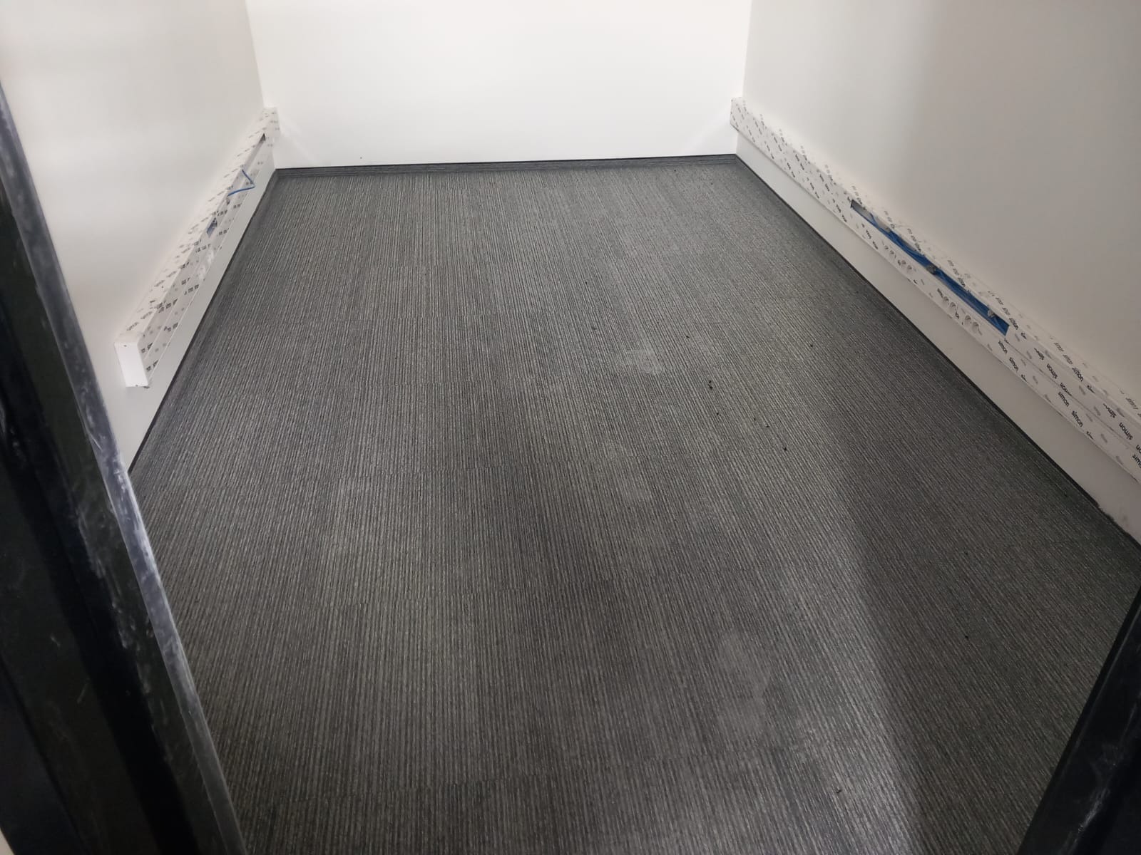 Szara wykładzina dywanowa w wąskim korytarzu, listwy przypodłogowe w folii ochronnej, widok z góry.