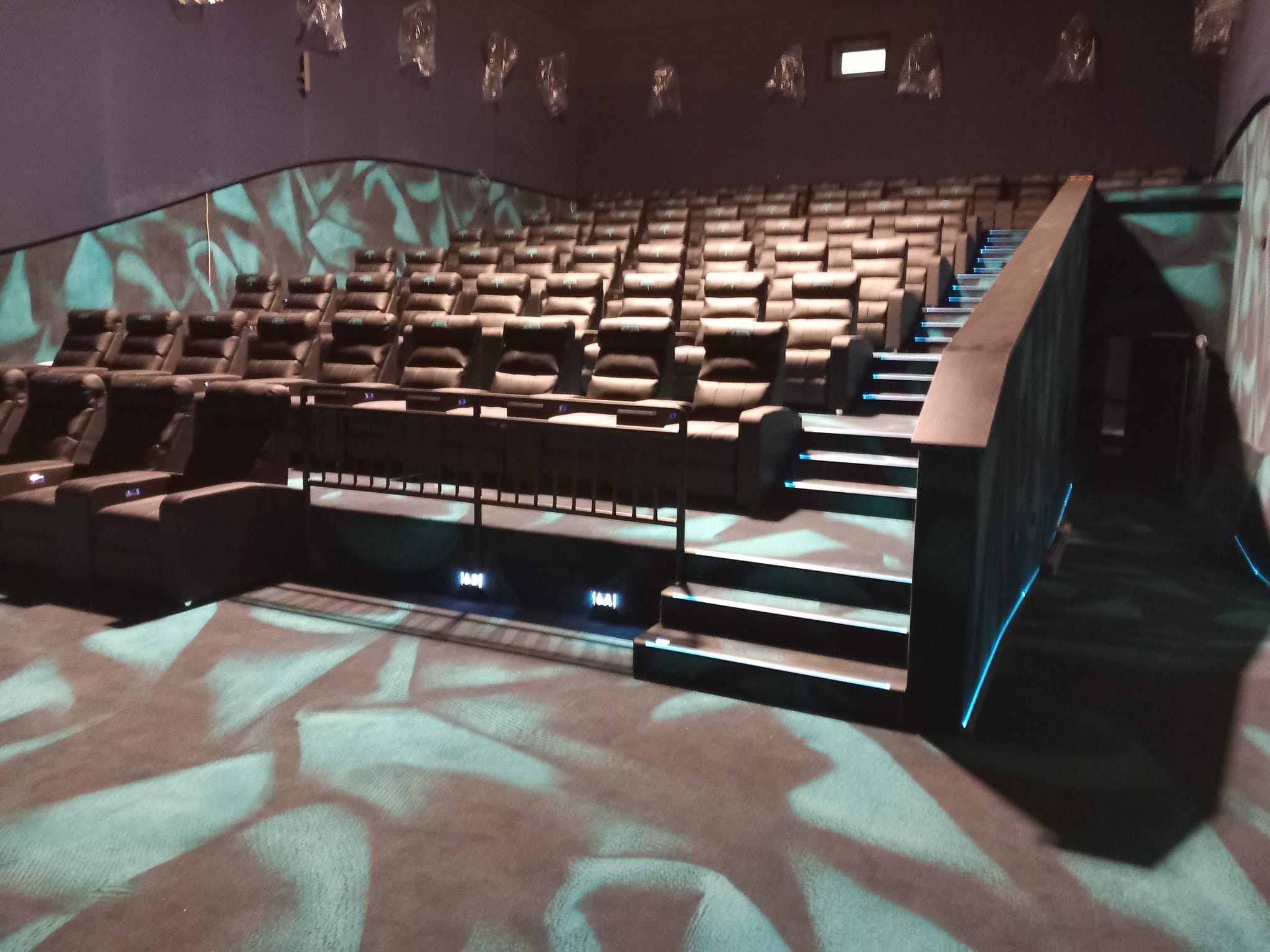 Sala kinowa z rzędami czarnych, skórzanych foteli, podświetlanymi schodami i wzorzystą wykładziną podłogową w odcieniach szarości i turkusu.