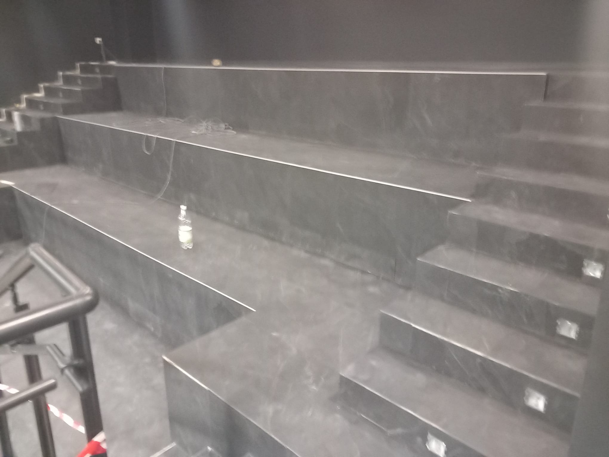 Schody wyłożone ciemnymi płytkami z metalowymi listwami, widoczne oświetlenie schodowe i butelka wody na jednym z stopni.