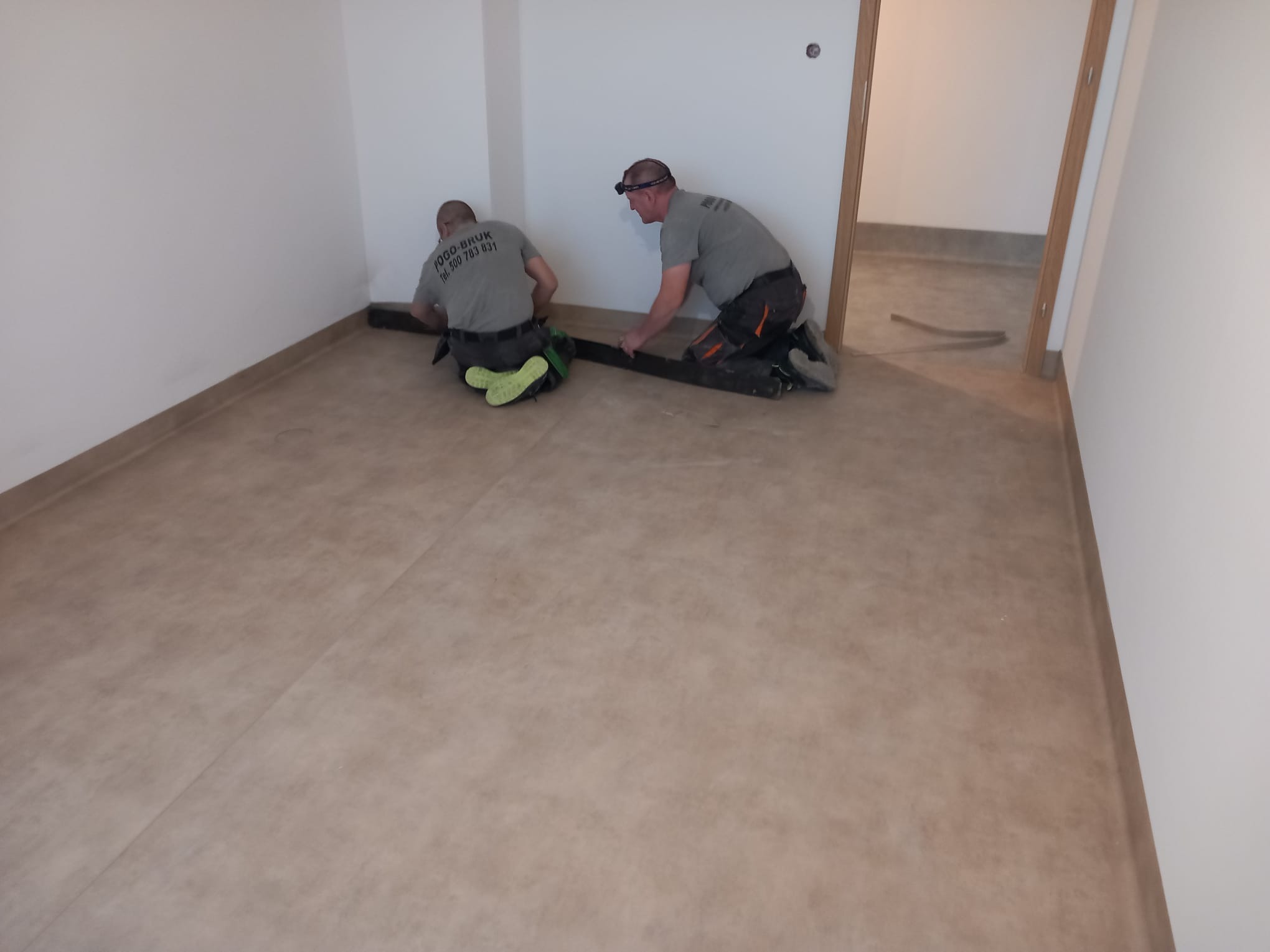 Dwóch monterów w szarych koszulkach z logo firmy 'POGO-BRUK' układa brązową wykładzinę w pustym pomieszczeniu, jeden z nich ma na głowie czołówkę.