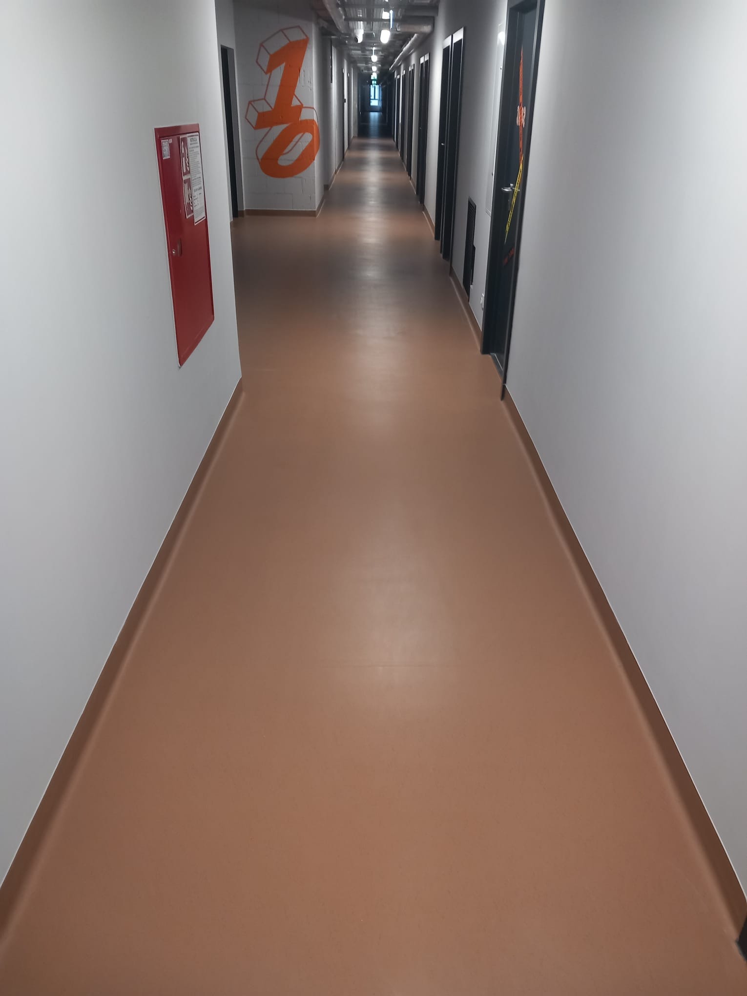 Długi korytarz z brązową, jednolitą podłogą, białymi ścianami i czarnymi drzwiami. Na ścianie widoczny czerwony hydrant i pomarańczowe graffiti z numerem.